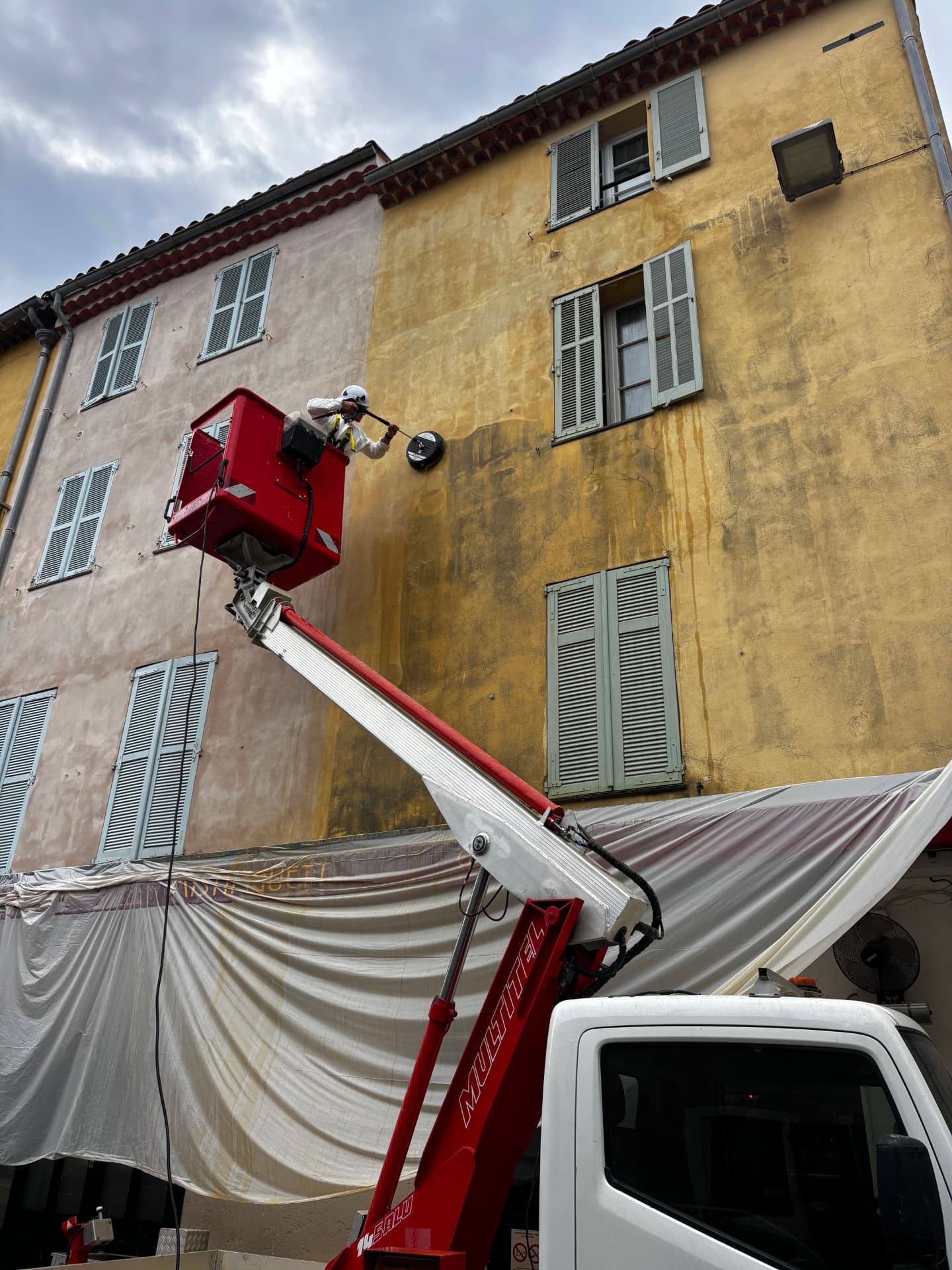 Technicien en nacelle nettoyant une façade ocre à la brosse rotative, bâches de protection posées en contrebas